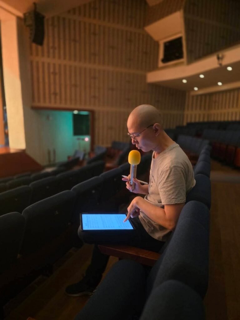 Composer Bang-hau Liao speaking at concert hall rehearsal holding a microphone and digital tablet